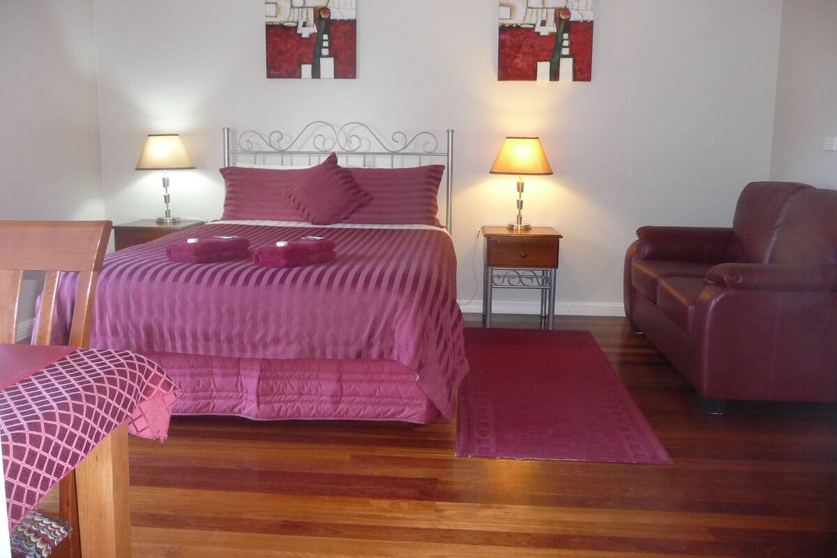 Quarto com cama, duas luminárias de cabeceira, mesa de cabeceira de madeira, poltrona de couro vermelha e duas pinturas abstratas na parede.