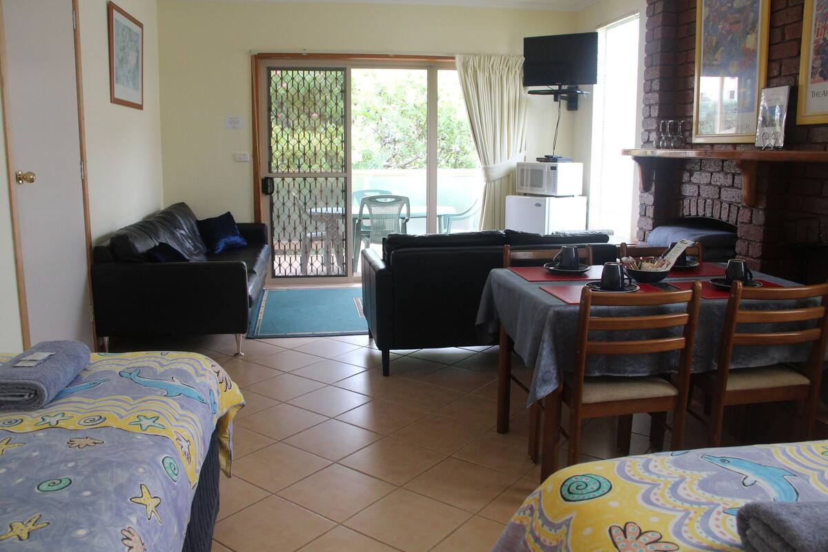 Quarto de hotel com duas camas, uma mesa de jantar, cadeiras, uma televisão e uma porta de vidro deslizante para o exterior.