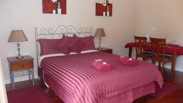 A neatly arranged bedroom with a large bed, two bedside tables with lamps, a wooden chair, and a dining table with a red tablecloth.