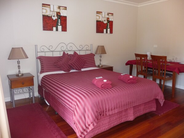 A neatly arranged bedroom with a large bed, two bedside tables with lamps, a wooden chair, and a dining table with a red tablecloth. - Angela's Beach Stays (Apollo Bay)