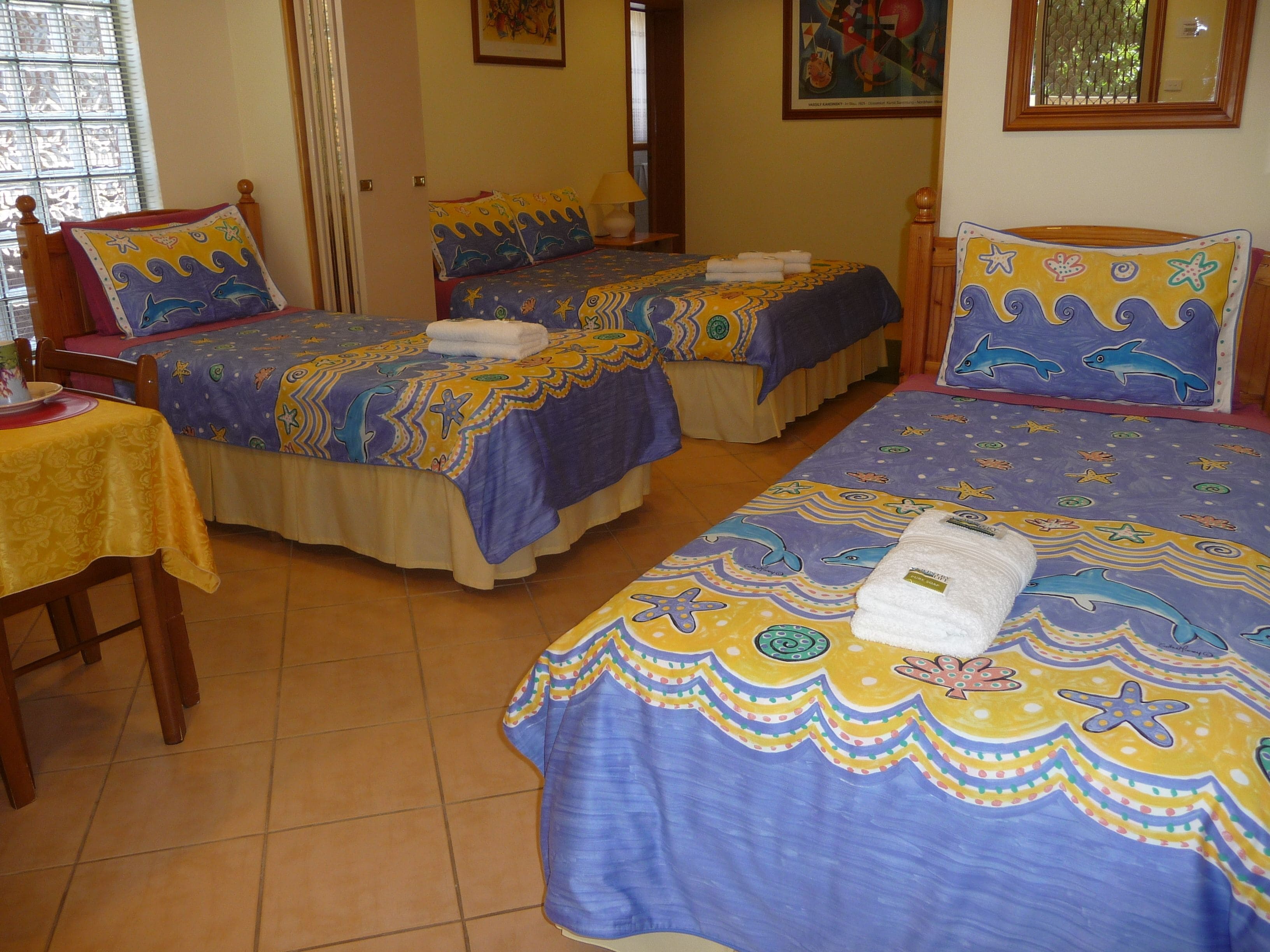 Quarto de hotel com duas camas, cada uma com colcha colorida com desenhos de criaturas marinhas. Há uma pequena mesa com toalha de mesa amarela e uma cadeira.
