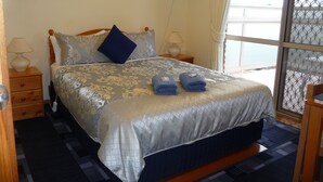 A neatly made bed with a patterned bedspread, two pillows, and two towels. There is a nightstand with a lamp, a framed picture on the wall, and a window with a view. - Angela's Beach Stays (Apollo Bay)