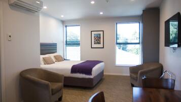 A bedroom with a bed, two armchairs, a wooden table, and a window with blinds.