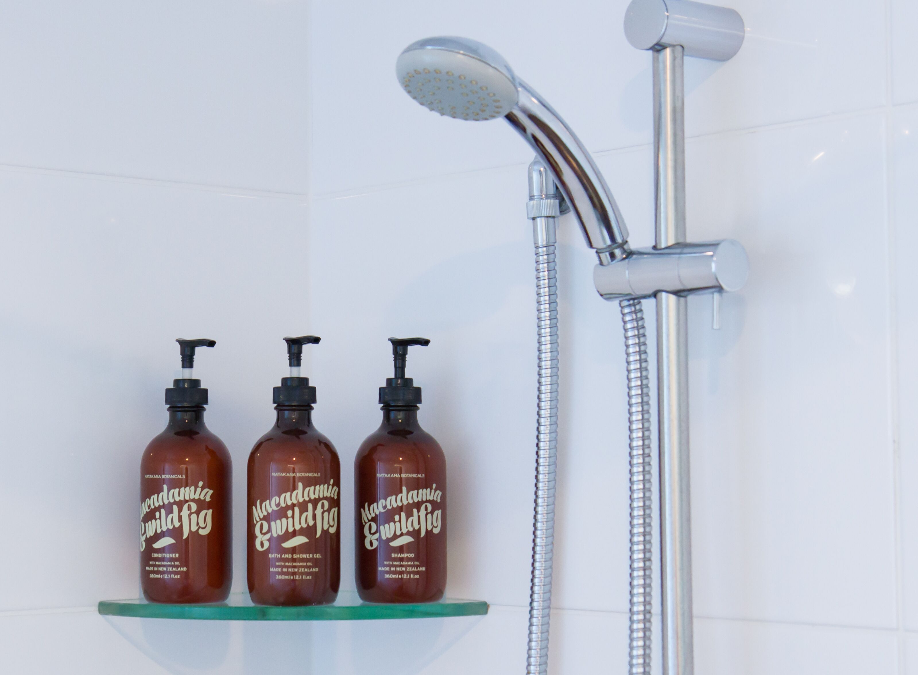 Three bottles of Macadamia Wild Fig shampoo and conditioner on a glass shelf next to a shower with a handheld showerhead.