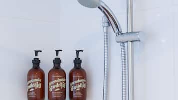 Tres botellas de champú y acondicionador Macadamia Wild Fig sobre un estante de vidrio junto a una ducha con cabezal de ducha manual.