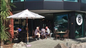 An outdoor seating area with tables and chairs, a white umbrella, and a glass storefront with a target logo. - 850 Cameron Motel (Tauranga)
