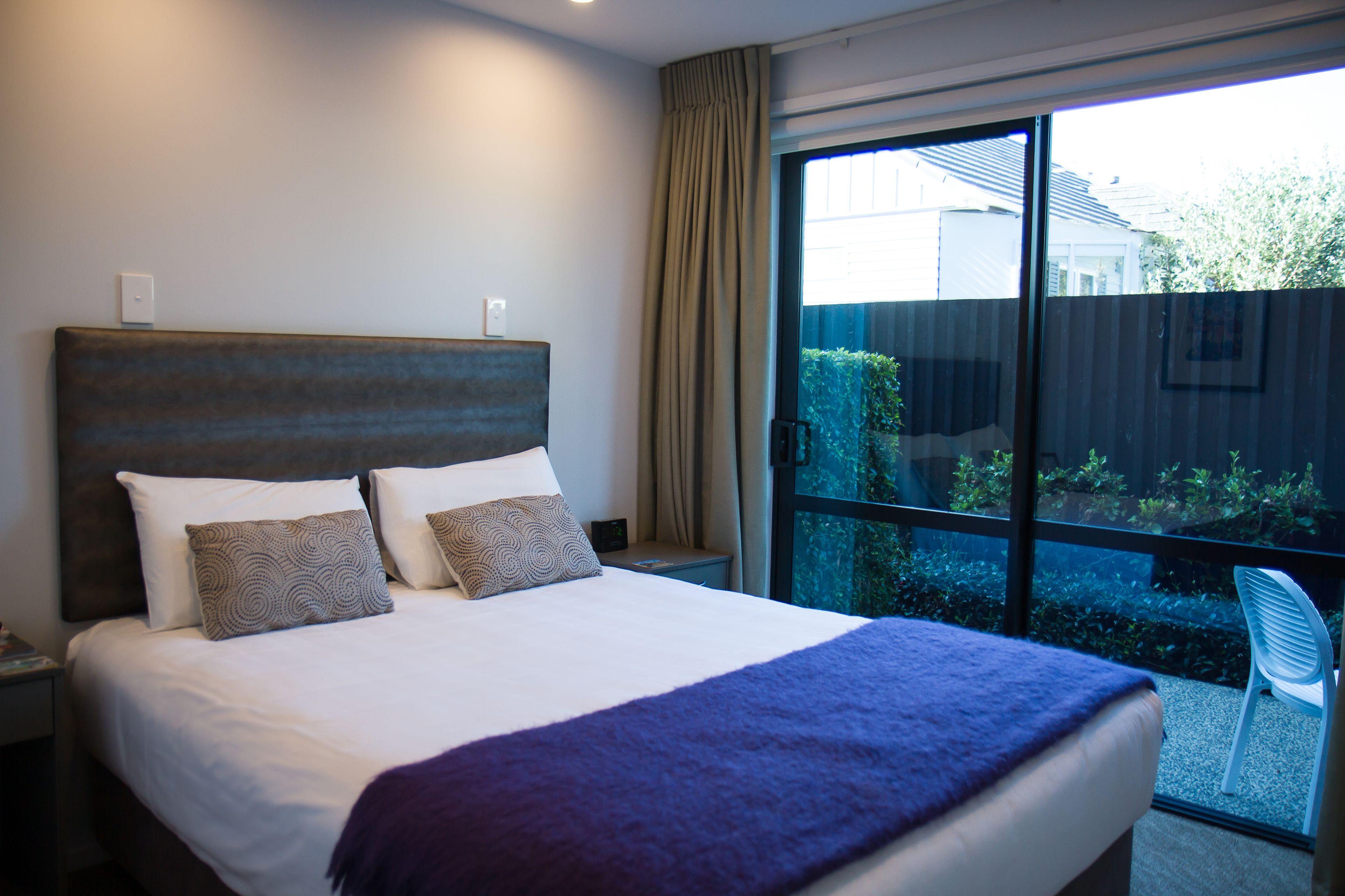 A hotel room with a large bed, a nightstand, a window with a view of greenery, and a sliding door to a balcony.