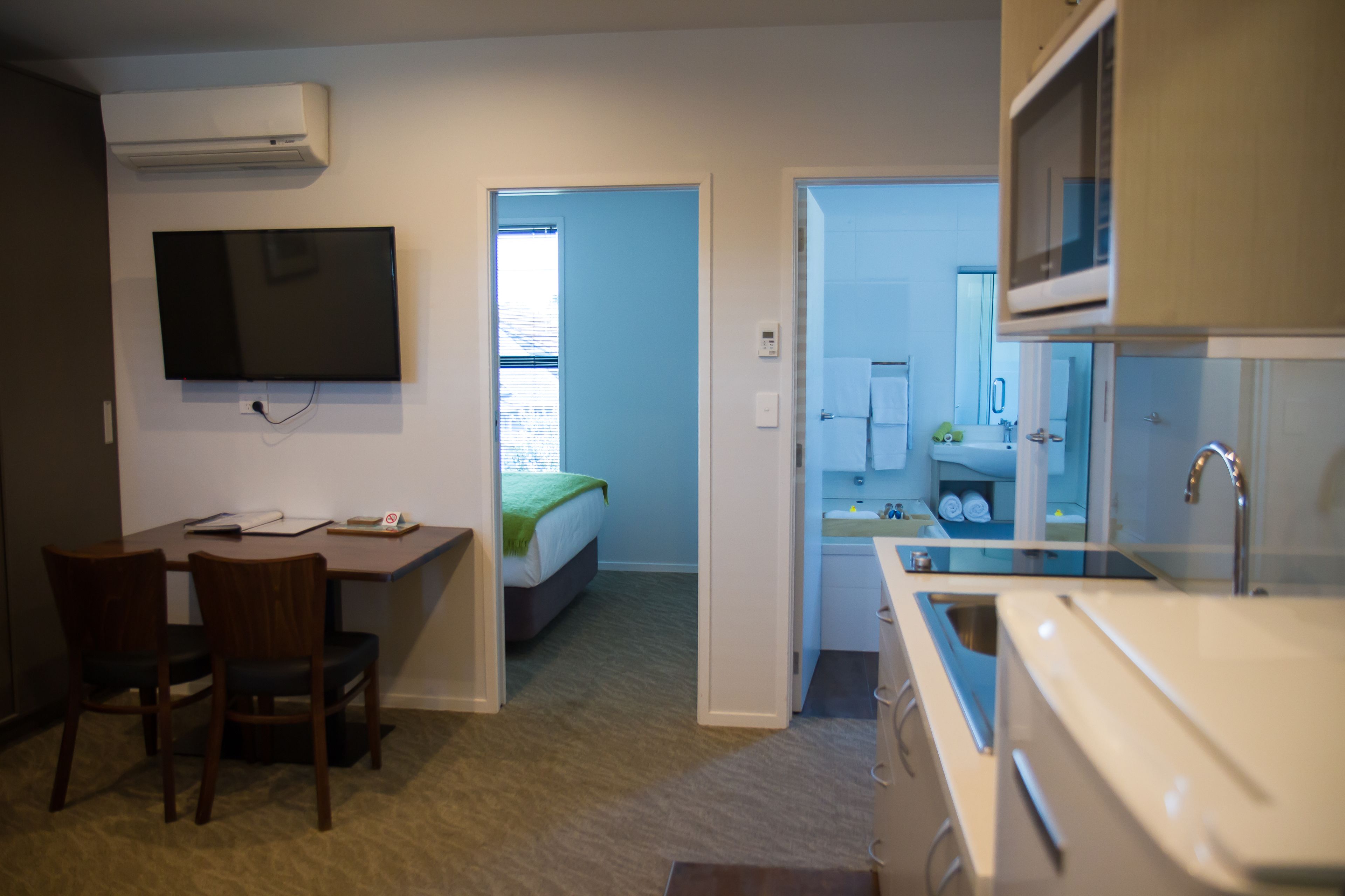 A compact room with a bed, a desk with chairs, a TV, and a kitchenette.