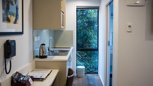A compact kitchen with a sink, kettle, and a small trash bin. There's a chair and a desk with a phone and a box of tissues. A window leads to a lush green outdoor area. - 850 Cameron Motel (Tauranga)
