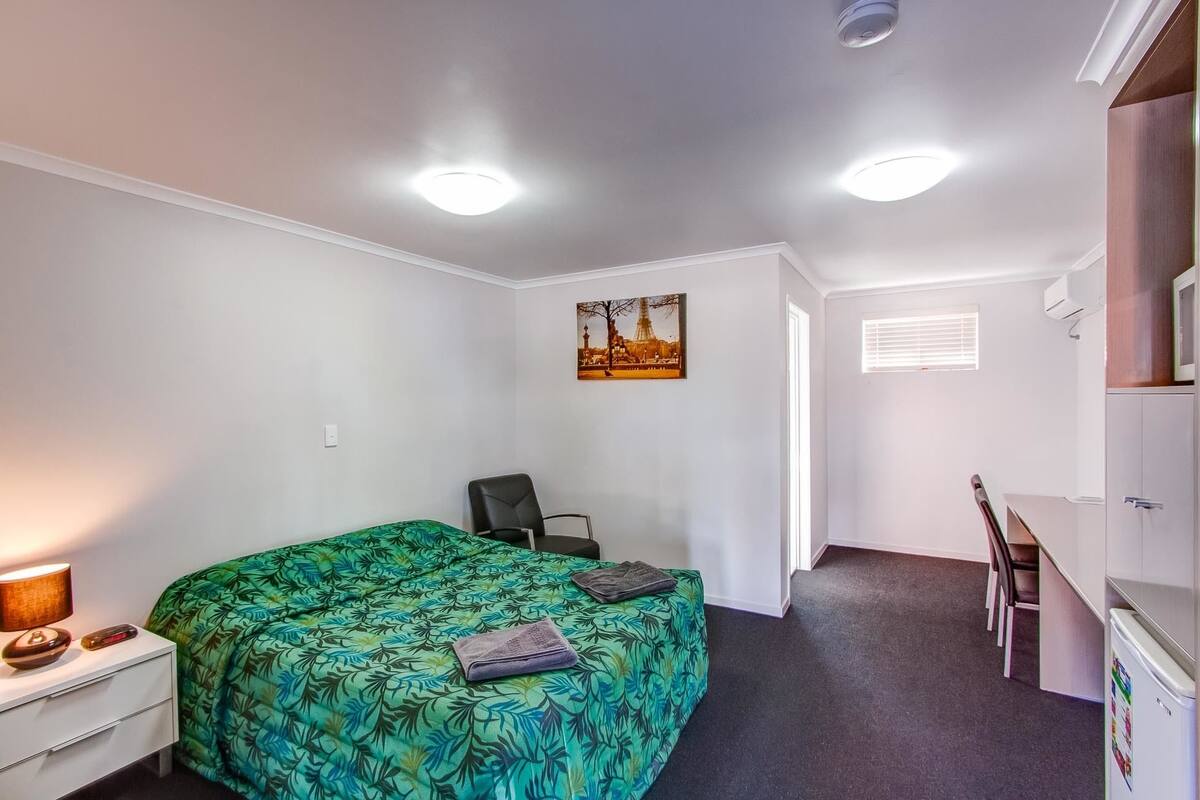 A bedroom with a bed, a desk, a chair, a lamp, and a painting on the wall.