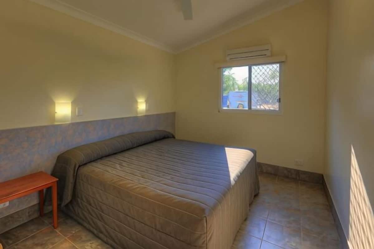 A bedroom with a bed, a nightstand, a window, and an air conditioning unit.