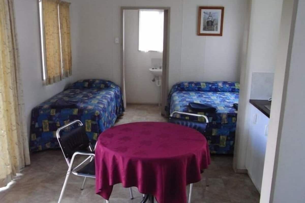 A small, compact room with two single beds, a table with a red tablecloth, a chair, and a sink in the corner.