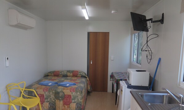 A compact room with a bed, a small table, a chair, a sink, and a television mounted on the wall.