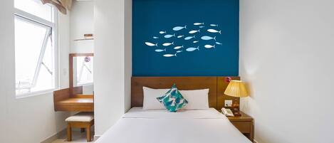 A hotel room with a large bed, a wooden headboard, a nightstand with a lamp, a window with curtains, and a wall decal of fish.
