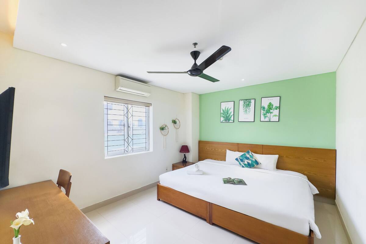 A hotel room with a bed, a desk, a chair, a ceiling fan, a window with bars, and wall-mounted plant illustrations.