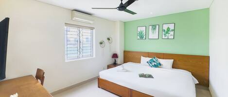 Une chambre d’hôtel comprenant un lit, un bureau, une chaise, un ventilateur de plafond, une fenêtre avec des barreaux et des illustrations de plantes accrochées au mur.