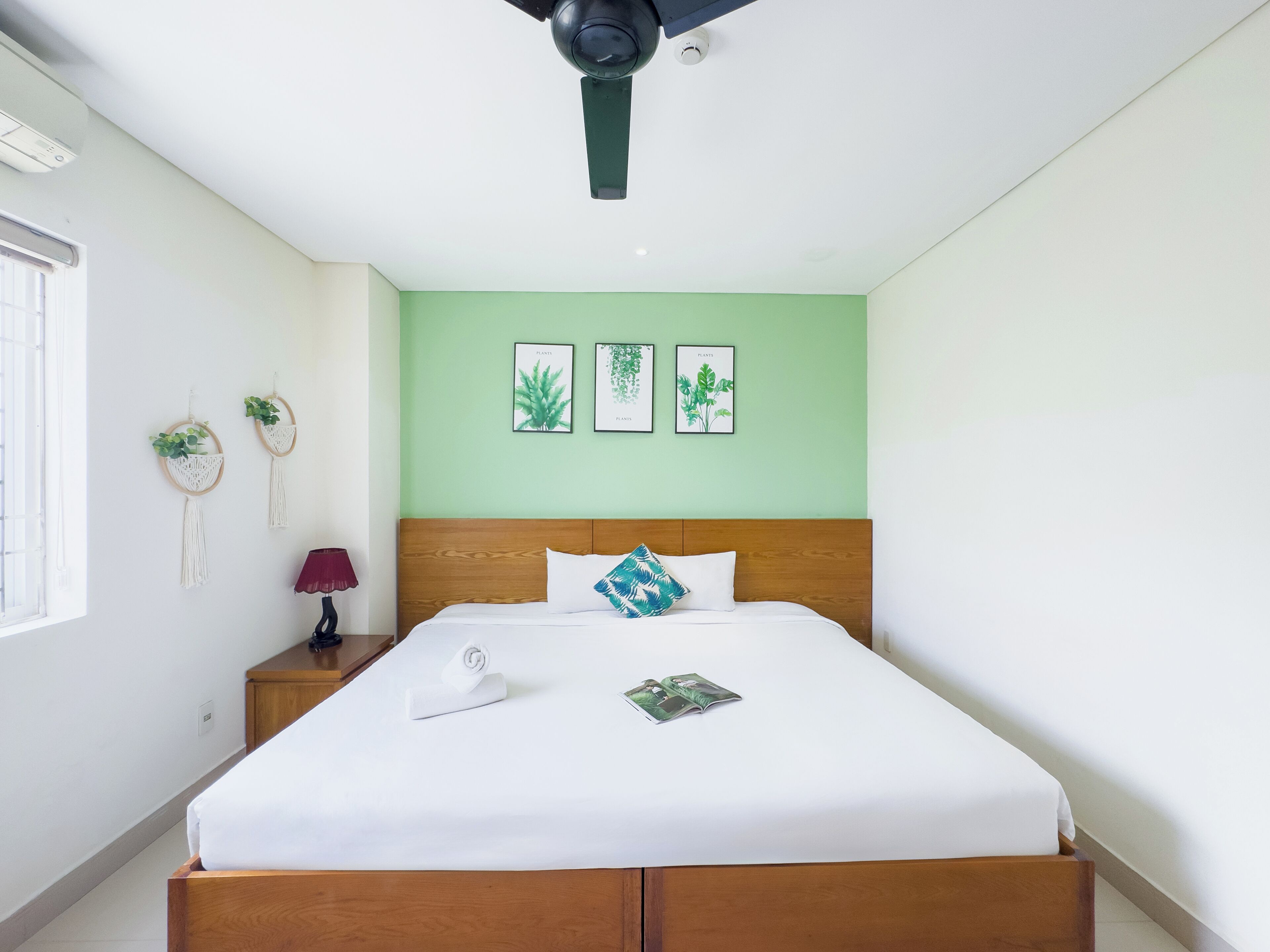 Una cama bien hecha con sábanas blancas, cabecera de madera y un ventilador de techo. Hay tres láminas enmarcadas con motivos botánicos en la pared sobre la cama.