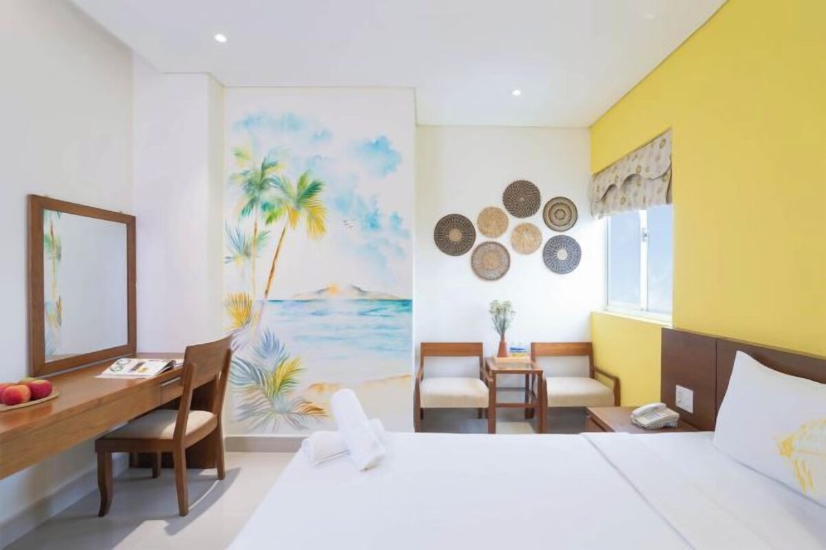 A hotel room with a bed, two chairs, a desk, and a painting of a beach scene.