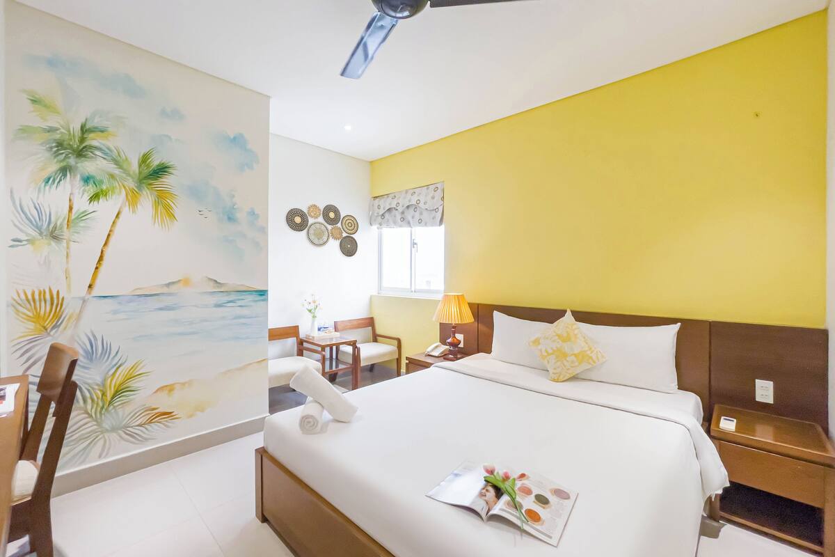 A hotel room with a bed, a desk, a chair, a wall with a tropical mural, and a ceiling fan.