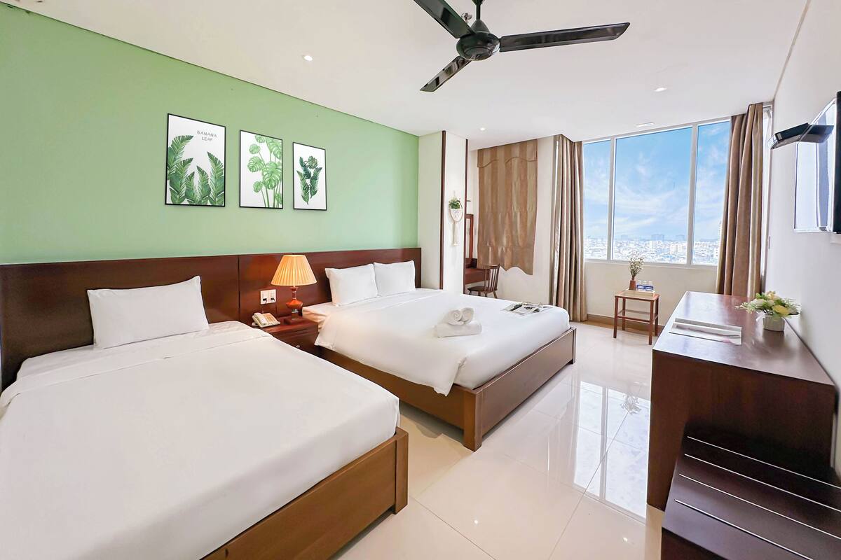 A hotel room with two beds, a desk, a chair, a ceiling fan, and large windows offering a city view.