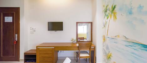 Una habitación de hotel con un escritorio de madera, un televisor montado en la pared, un ventilador de techo y un mural con motivos playeros.