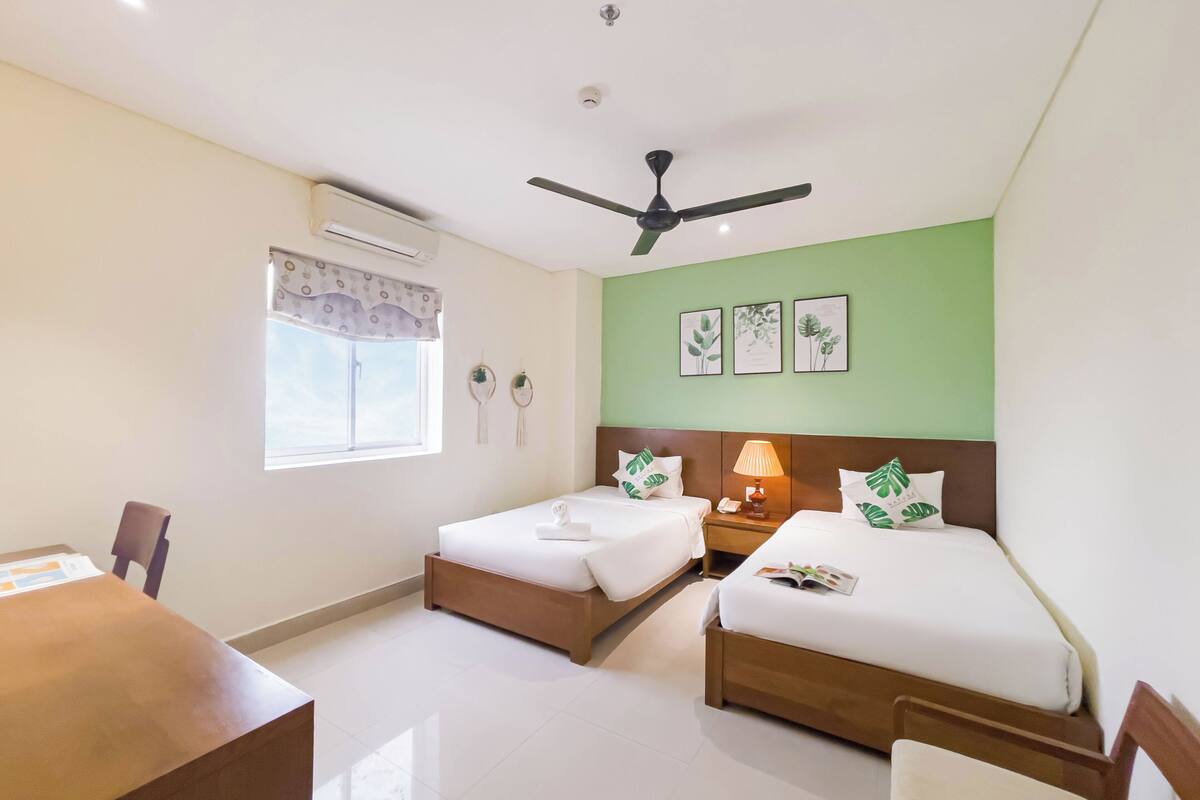 A hotel room with two beds, a desk, a chair, a ceiling fan, and a window with a curtain.