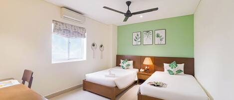 A hotel room with two beds, a desk, a chair, a ceiling fan, and a window with a curtain.