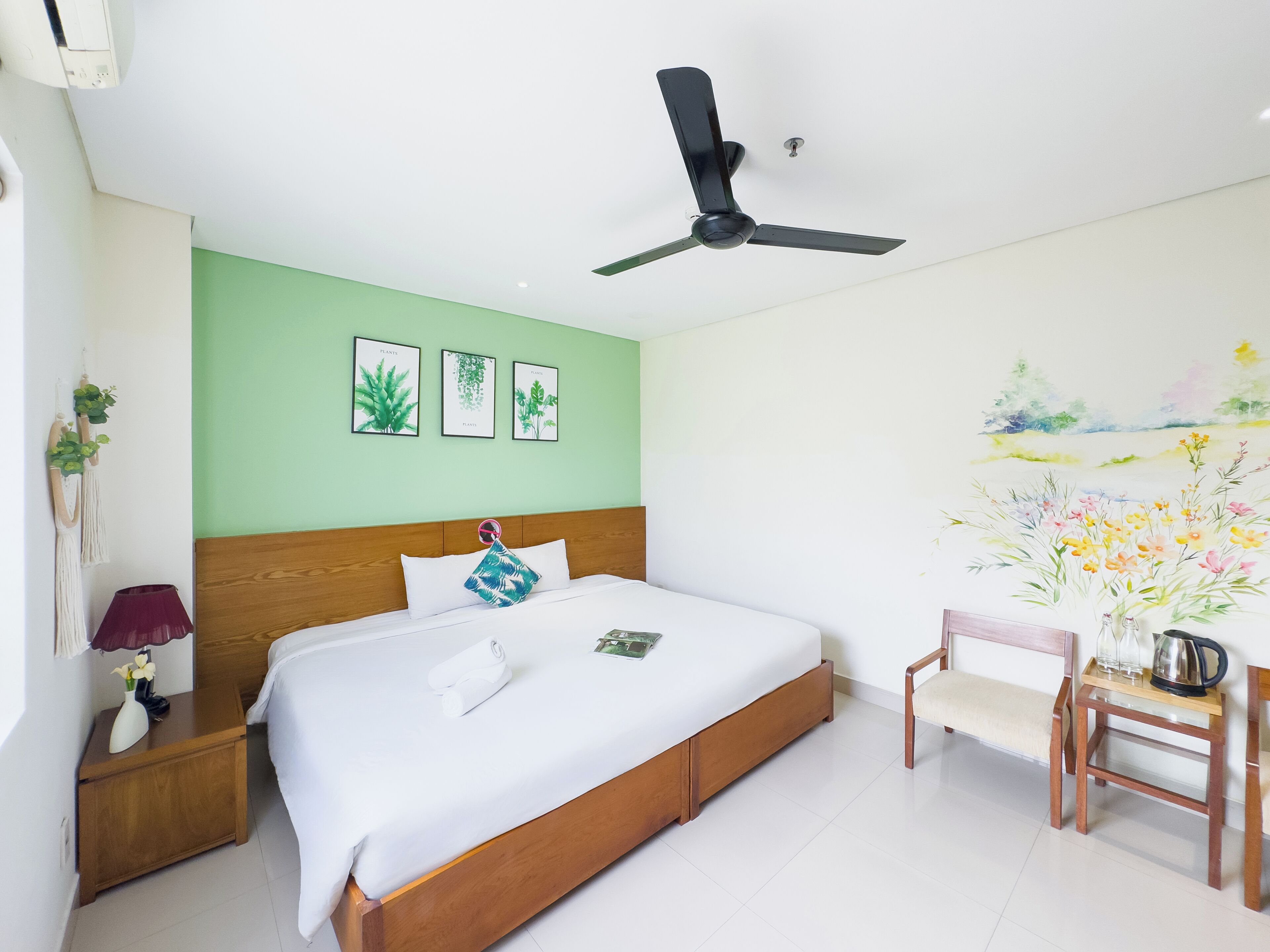 Une chambre à coucher avec un lit, un ventilateur de plafond, une table de chevet avec une lampe, une chaise et un mur décoré d’illustrations de plantes.