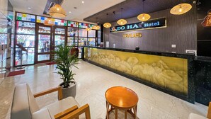 Un espace d’accueil d’hôtel au design moderne, comprenant un comptoir d’accueil, un coin salon et des éléments décoratifs tels que des plantes et des œuvres d’art mural.