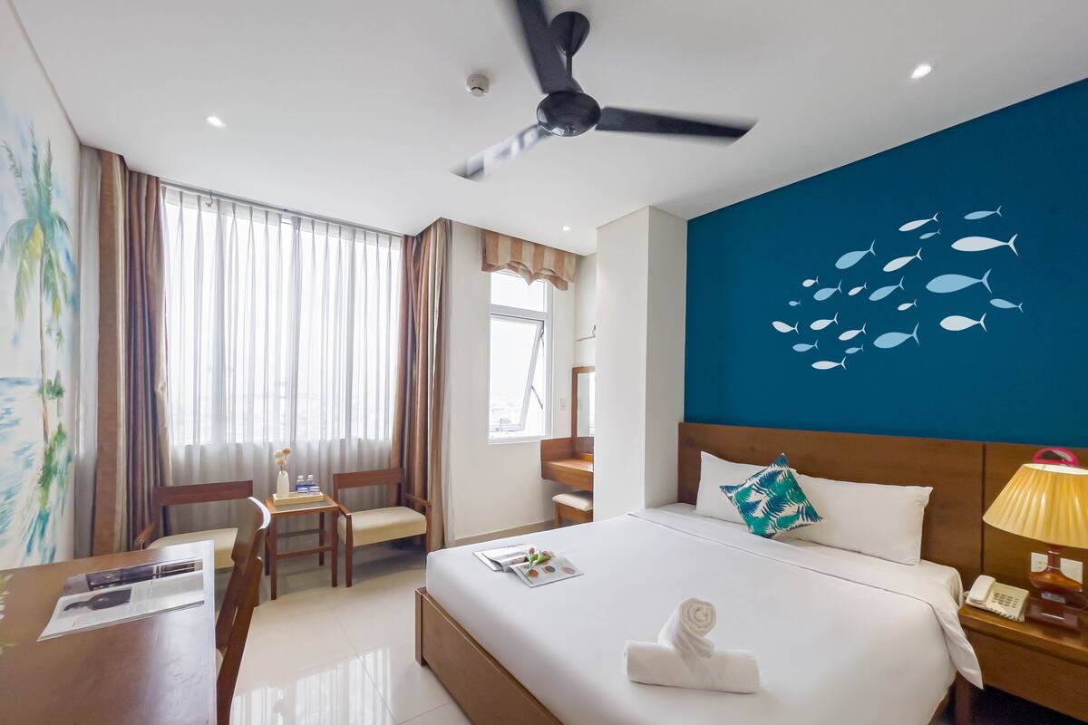 A hotel room with a bed, a desk, a chair, a ceiling fan, and a wall with a fish mural.