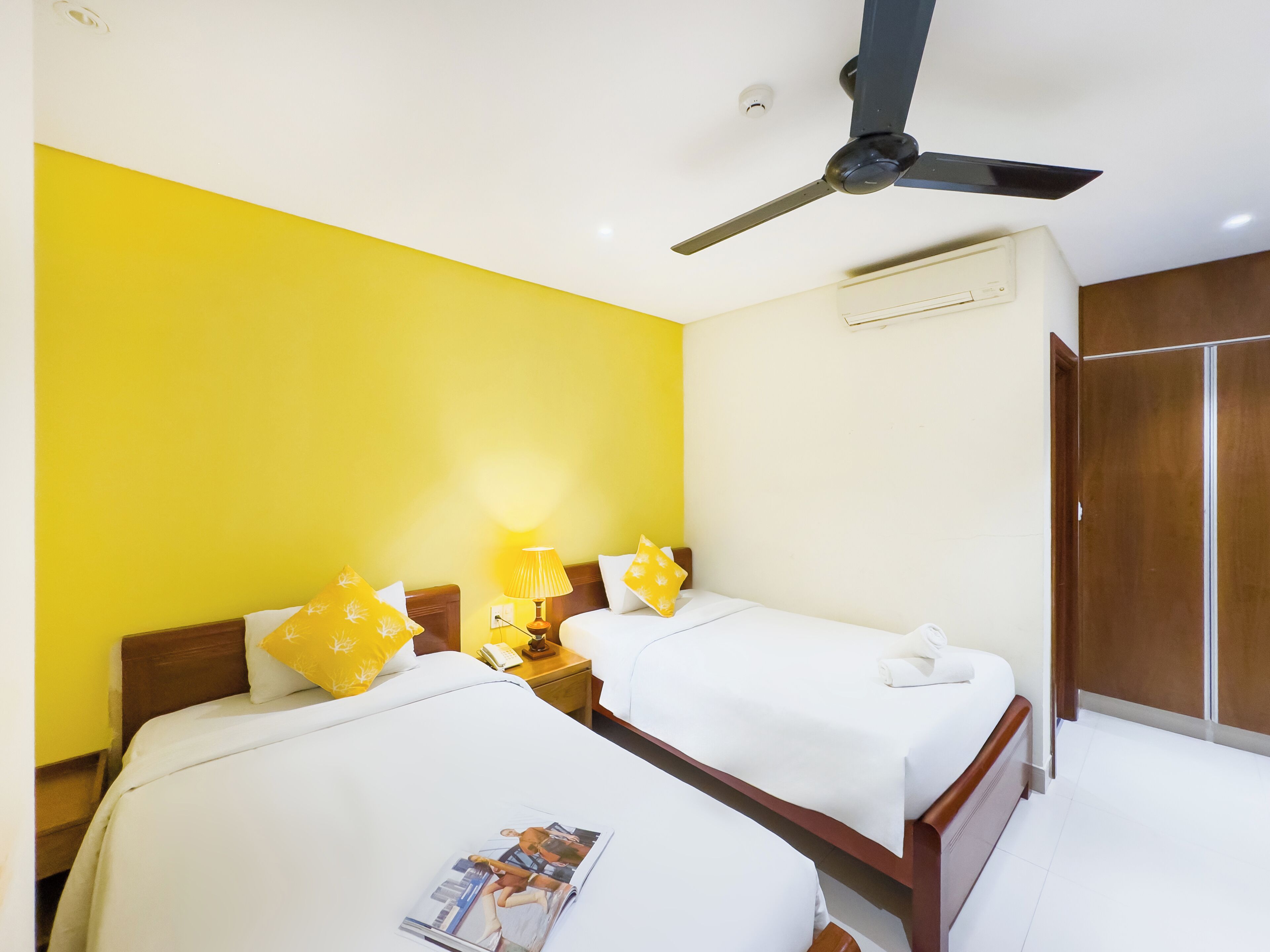 A hotel room with two beds, a ceiling fan, a nightstand with a lamp, and a wardrobe.