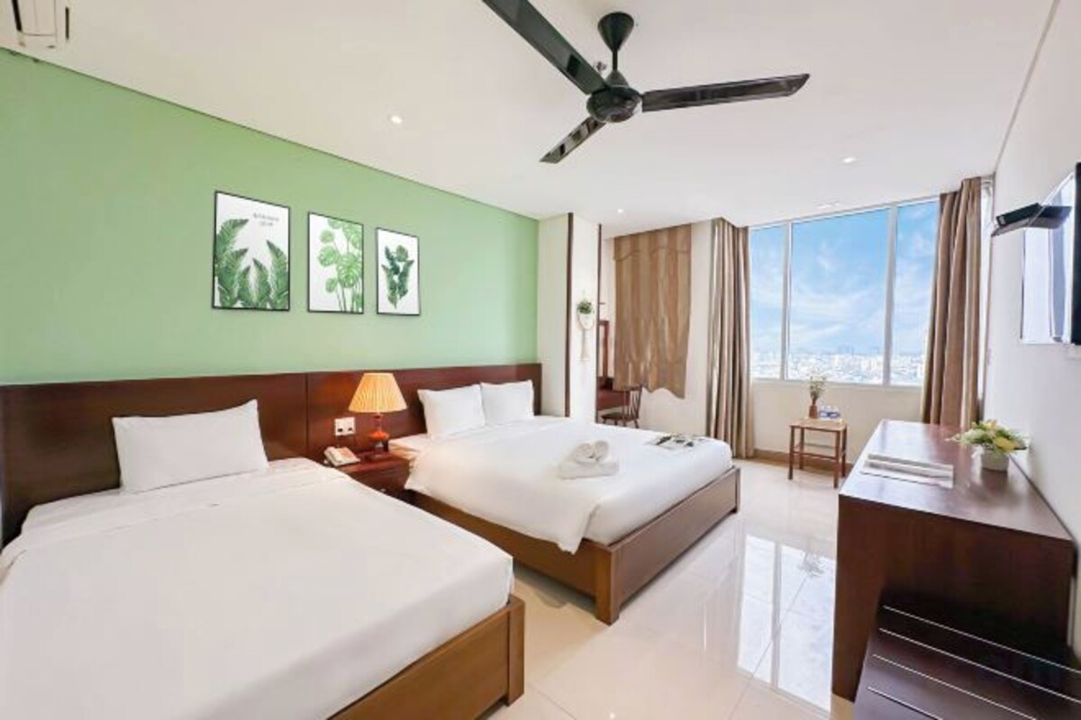 A hotel room with two beds, a desk, a chair, a ceiling fan, and a large window with a city view.