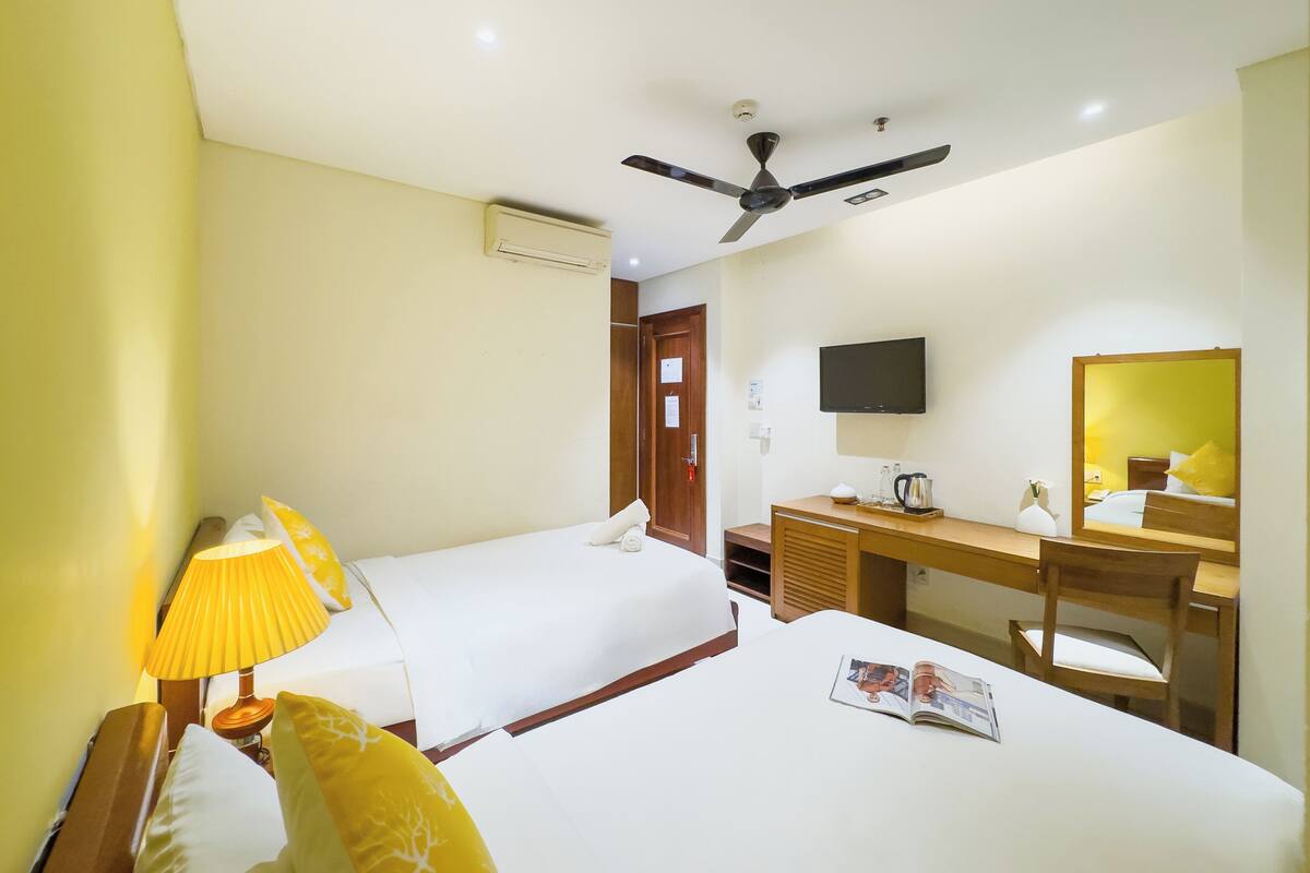 A hotel room with two single beds, a desk with a chair, a television, and a ceiling fan.