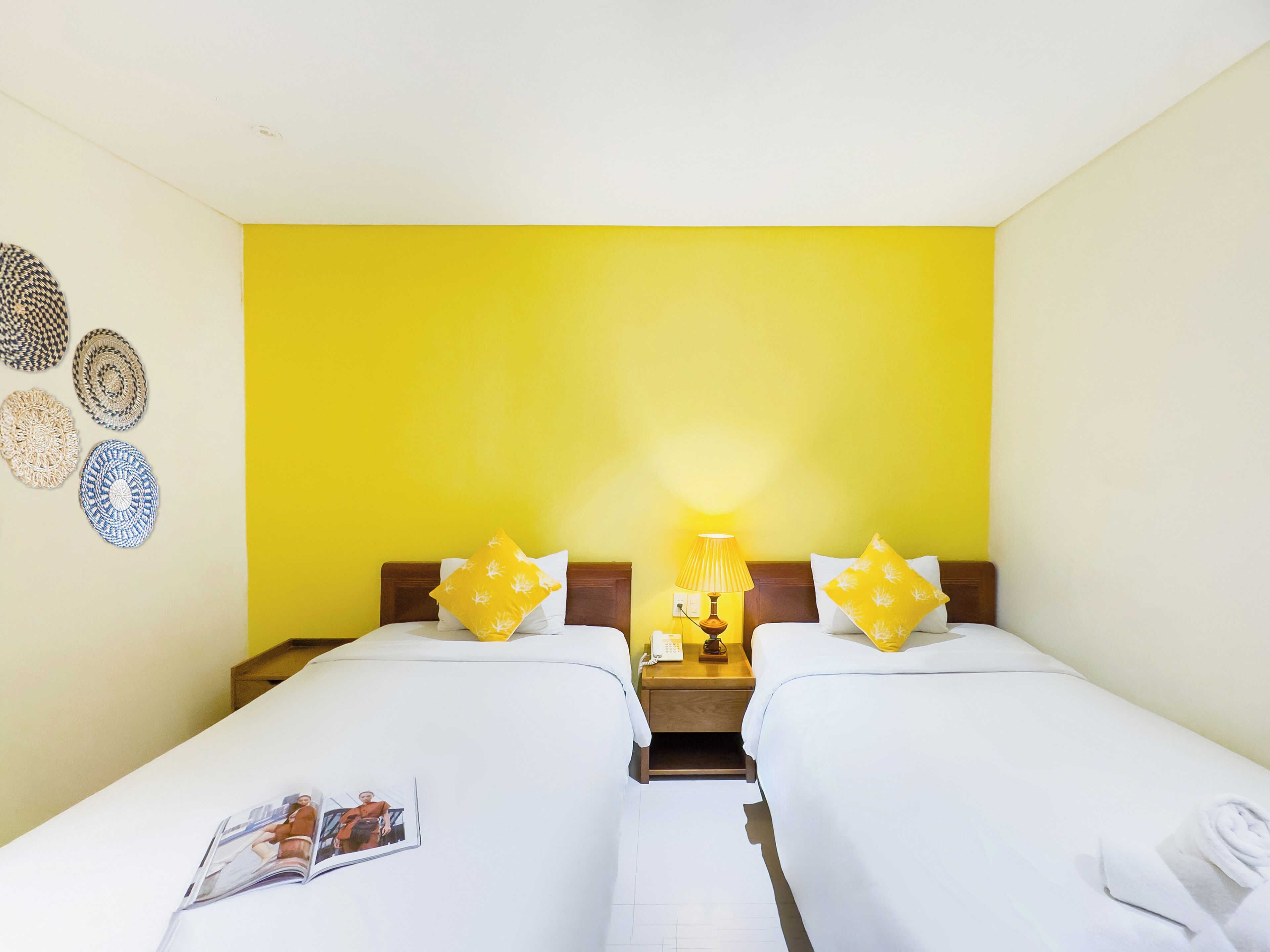 A hotel room with two beds, a wooden nightstand with a lamp, and decorative wall hangings.
