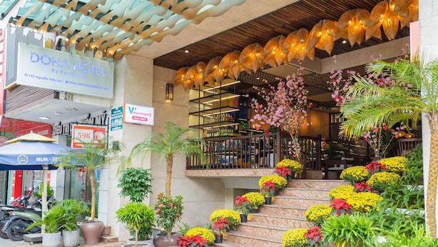 L’extérieur de l’HÔTEL DOHA, de la chaîne Merrelland, se distingue par un escalier orné de fleurs jaunes et de plantes en pot, une enseigne et un panneau « V+ » indiquant la présence d’un restaurant à proximité.