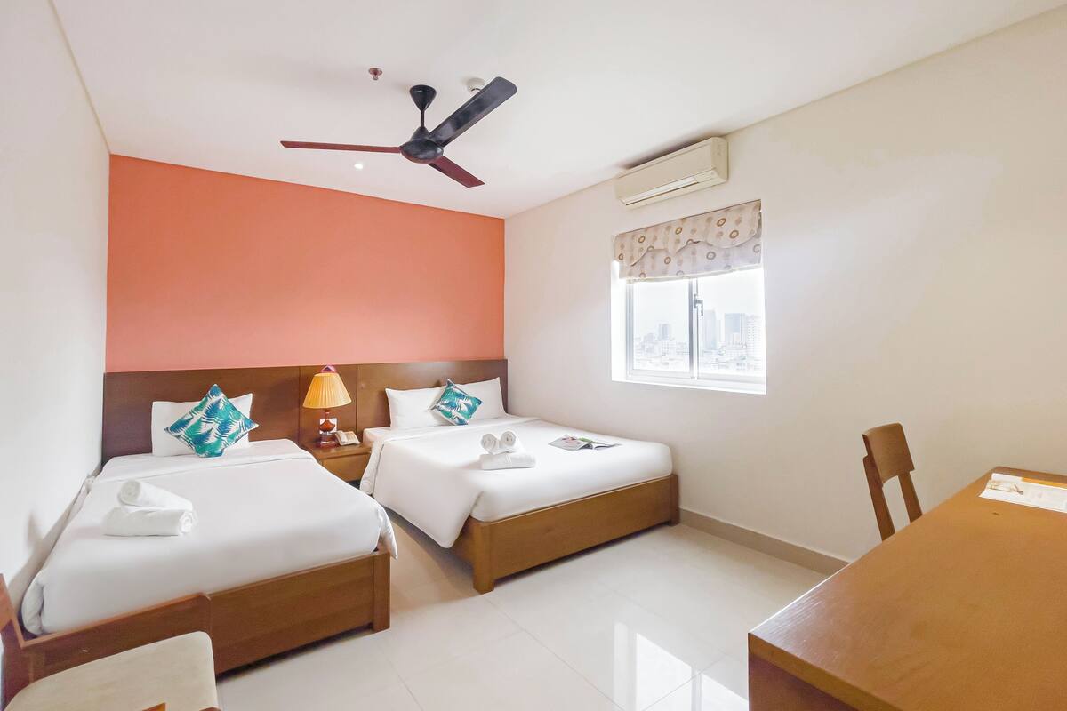 A hotel room with two beds, a ceiling fan, an air conditioning unit, a window with a view of the city, and a wooden table.