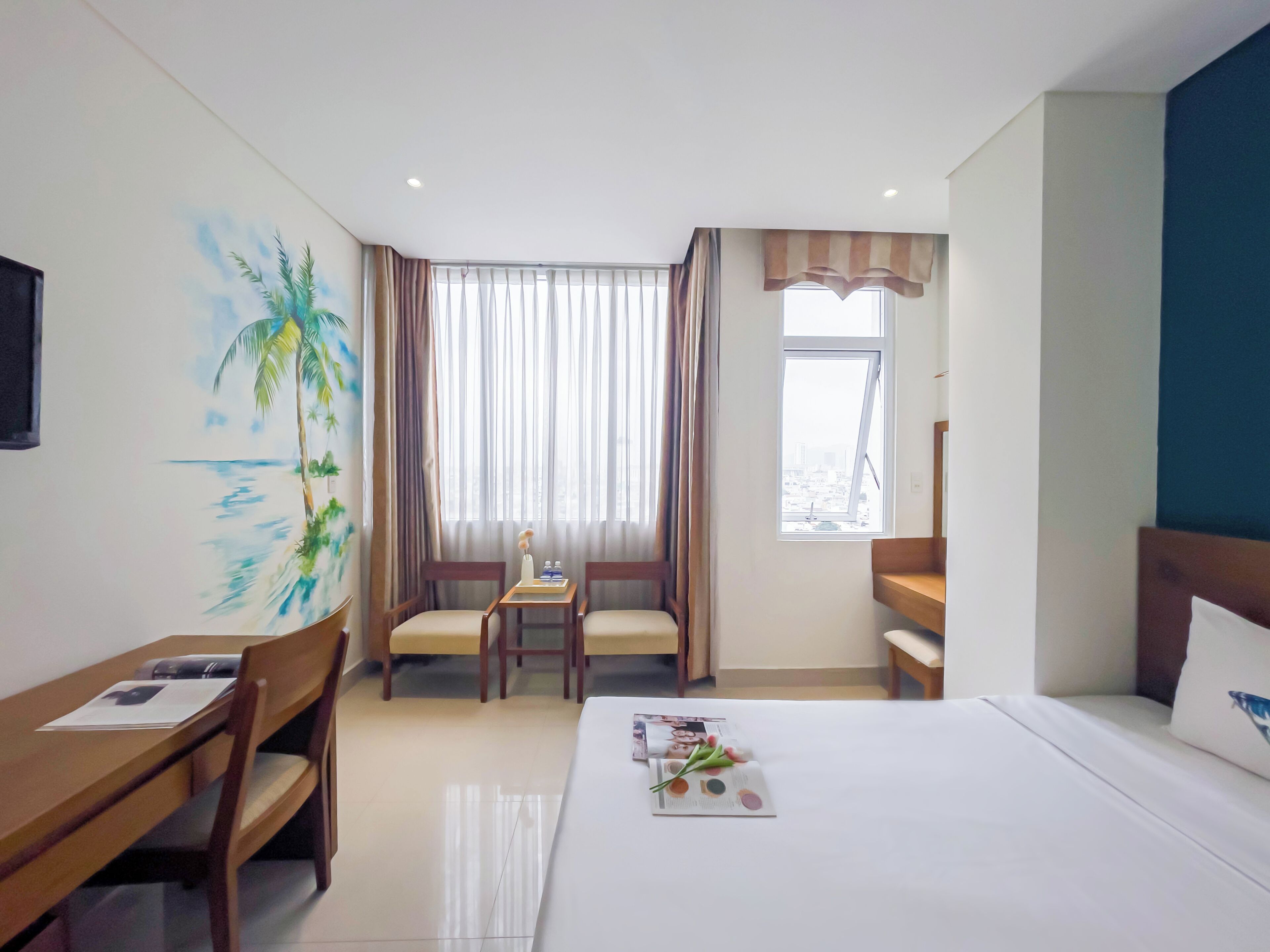 A hotel room with a bed, a desk with chairs, a painting of a beach scene, a window with curtains, and a view of the city.
