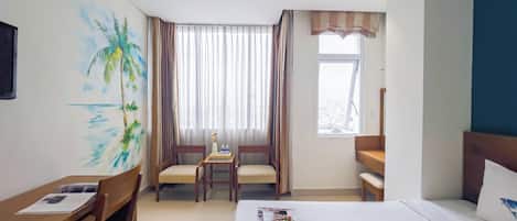 Una habitación de hotel con cama, escritorio con sillas, un cuadro de una playa y ventana con cortinas, además de vistas a la ciudad.