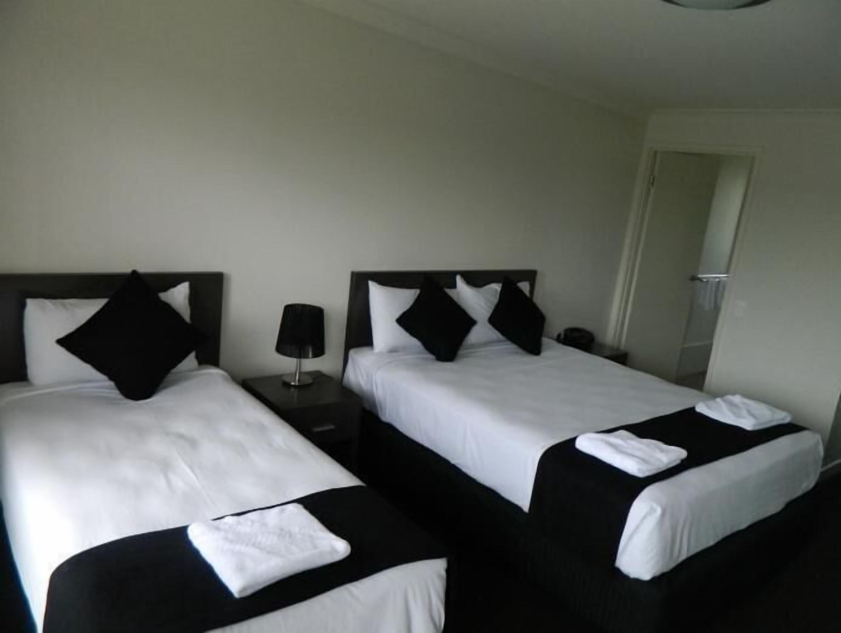 Una habitación de hotel con dos camas, cada una con ropa de cama blanco y negro, una mesita de noche con lámpara y una puerta que da a otra habitación.