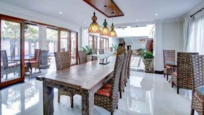 Un comedor amplio con una larga mesa de madera y sillas de mimbre, ventanas grandes y lámparas colgantes.
