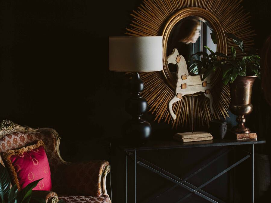 A dark room with a vintage chair, a mirror with a sunburst frame, and a lamp on a console table.