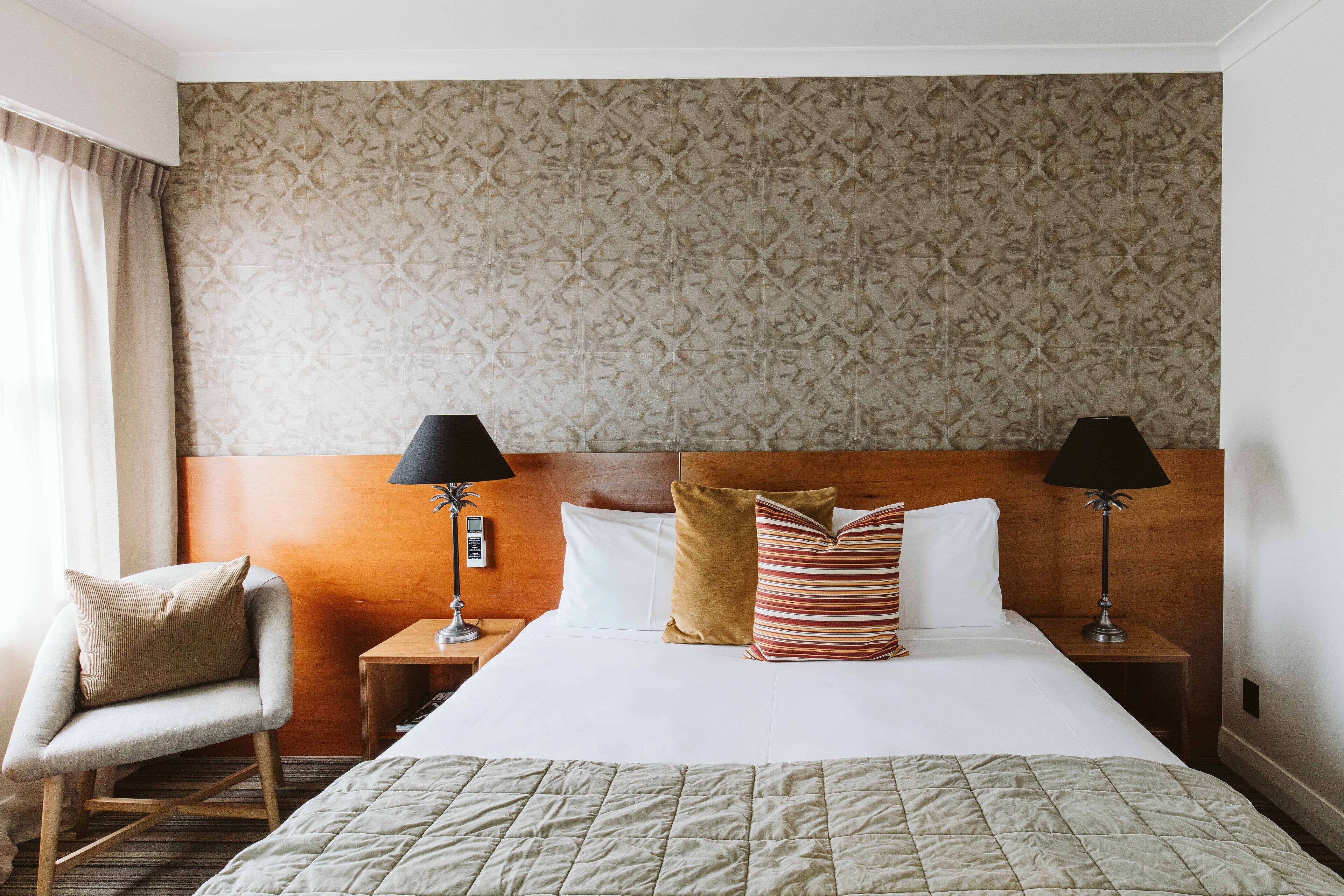 Ein Hotelzimmer mit einem großen Bett, zwei Nachttischen mit Lampen, einem Sessel und einer Wand mit Muster.