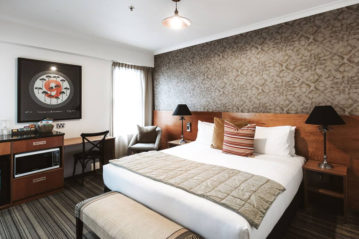 A hotel room with a large bed, two bedside tables with lamps, a desk with a chair, a microwave, and a framed artwork on the wall.