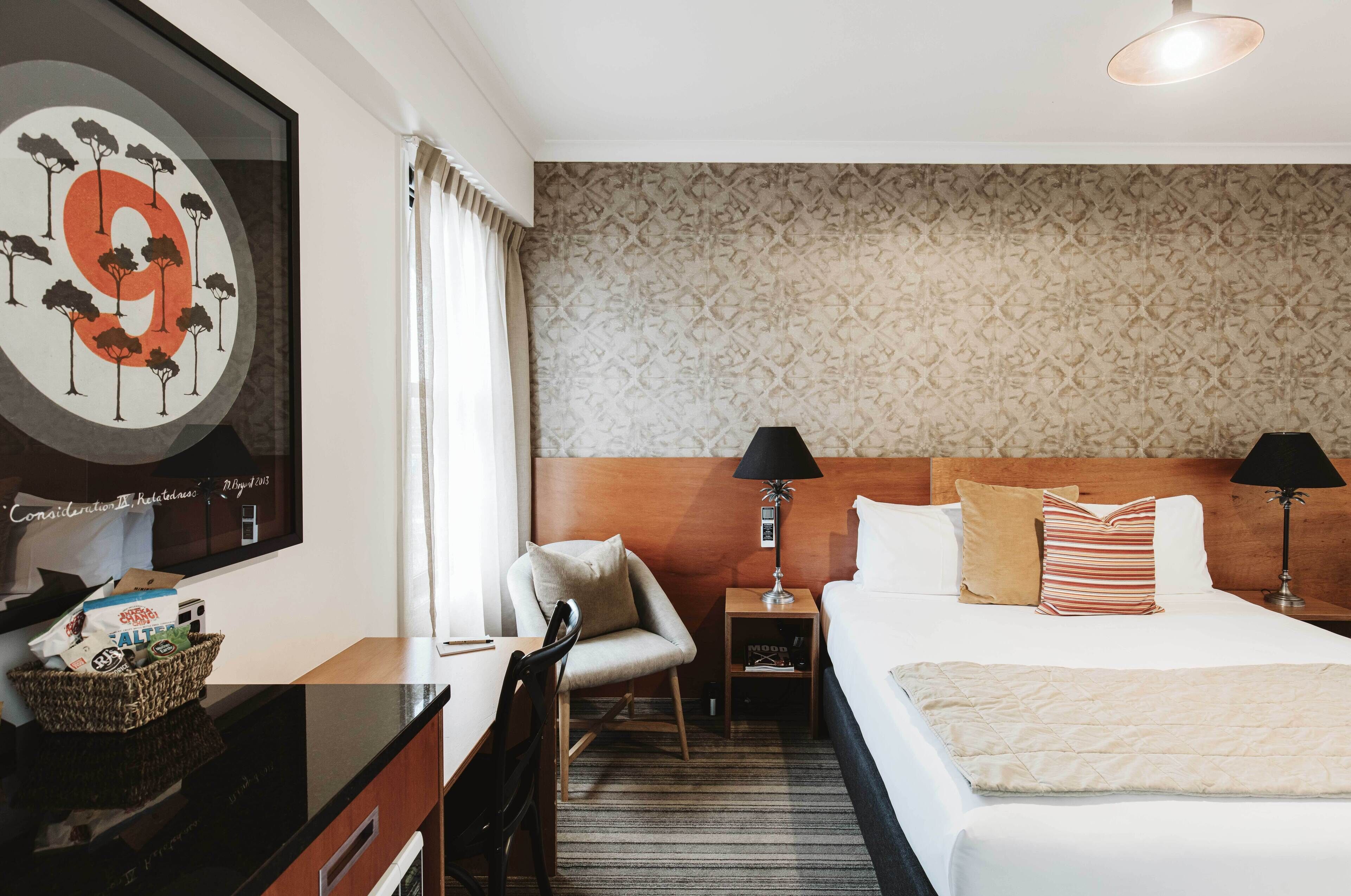 A hotel room with a large bed, two bedside tables with lamps, a chair, a desk with a basket of items, and a wall with a framed artwork.