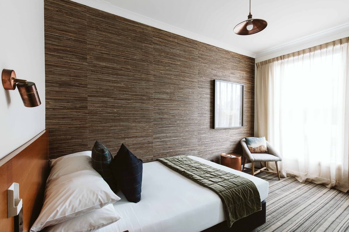 A modern hotel room with a wooden accent wall, a large bed with a green throw, a side table, a chair, and a large window with curtains.
