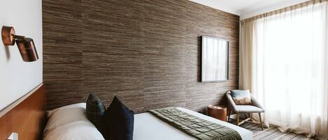 A modern hotel room with a wooden accent wall, a large bed with a green throw, a side table, a chair, and a large window with curtains.