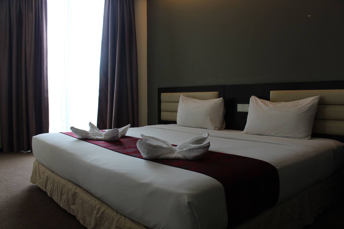 A hotel room with a large bed, two pillows, a red throw blanket, and a white towel arrangement on the bed.