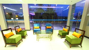 Un vestíbulo con cuatro sillones verdes y una mesa de centro de vidrio, amplias ventanas con vistas a palmeras y un estacionamiento, y un letrero de neón al fondo.