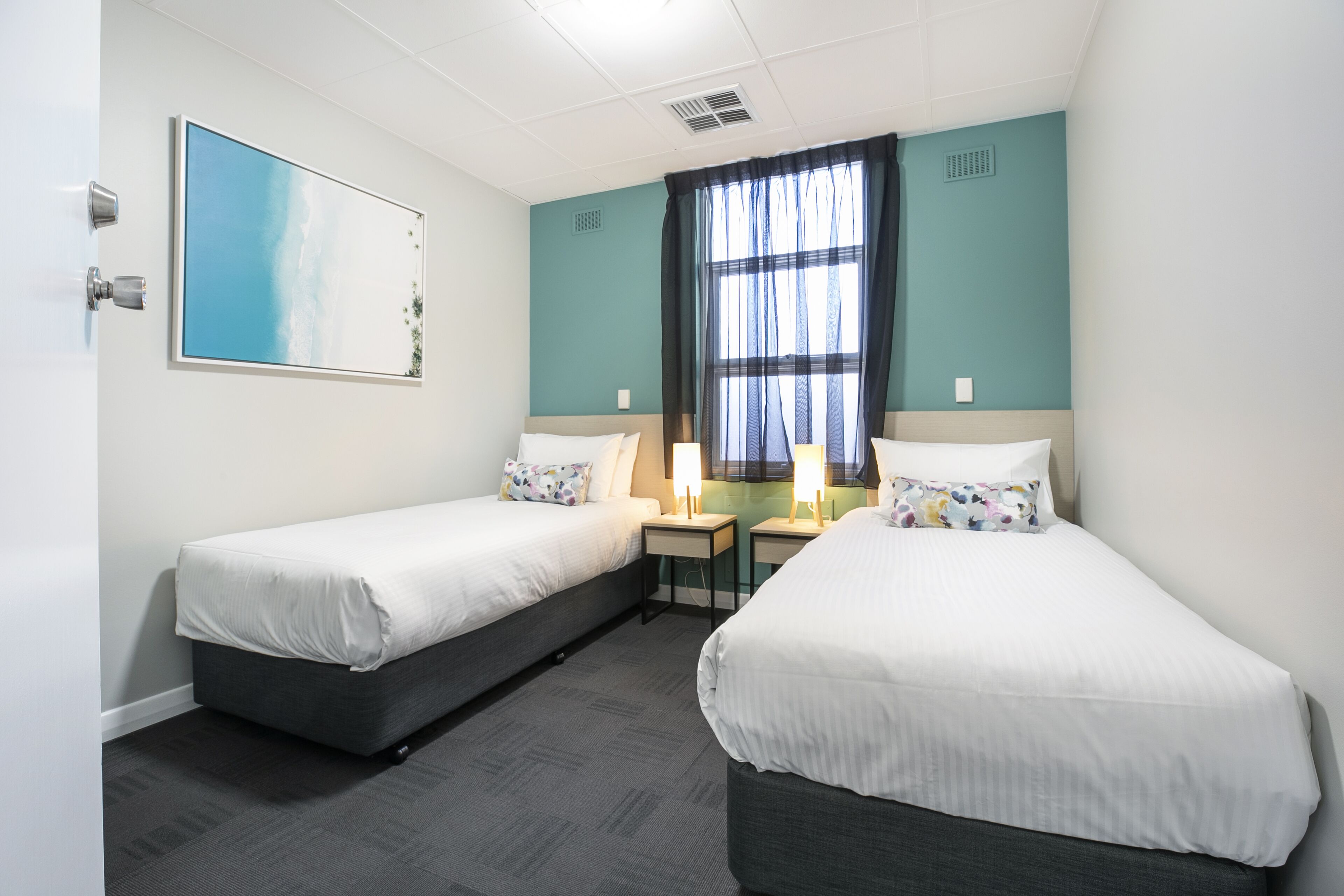 Een hotelkamer met twee eenpersoonsbedden, een groot raam met dunne gordijnen, een schilderij aan de muur en een kleine tafel met een lamp.