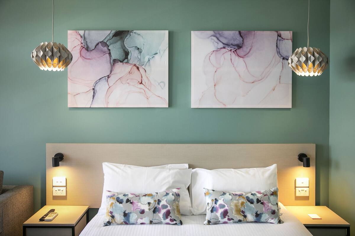 A hotel room with a bed, two wall-mounted paintings, and two pendant lights.